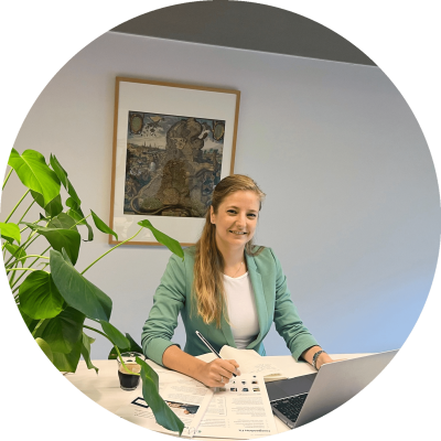 een vrouw zit aan een bureau met een laptop erop, en kijkt lachend in de camera
